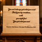A rectangular wooden plaque engraved with a motivational quote and songwriters' names, displayed on a dark wooden altar stand in a church setting with blurred stained glass and candles in the background.
