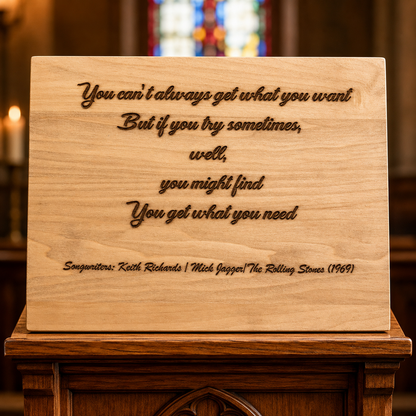 A rectangular wooden plaque engraved with a motivational quote and songwriters' names, displayed on a dark wooden altar stand in a church setting with blurred stained glass and candles in the background.